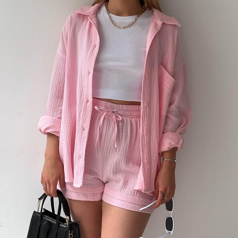 Frau in rosa Freizeitanzug mit Hemd und Shorts, hält schwarze Handtasche und weiße Sonnenbrille. Modetrend, Sommeroutfit, lässiger Stil.