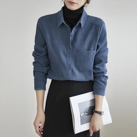 Frau in blauer Bluse mit Brusttasche, hält ein Magazin, trägt schwarzen Rock und Armbanduhr. Elegante, moderne Damenmode.
