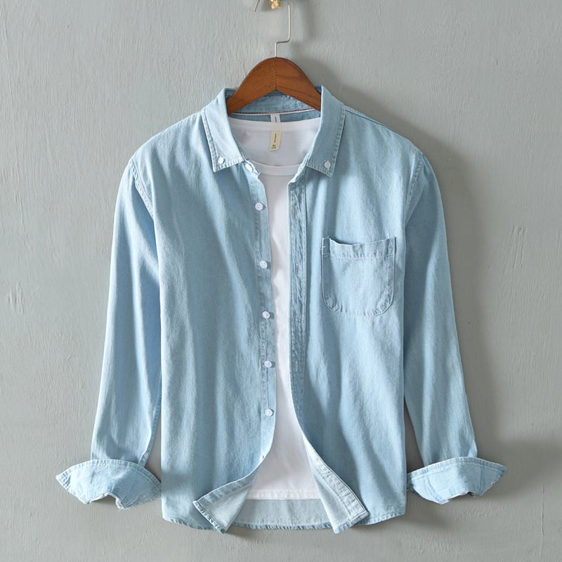 Hellblaues Jeanshemd mit Knopfleiste und Brusttasche, über weißem T-Shirt, auf Holzbügel vor grauer Wand. Modetrend, Herrenbekleidung, Freizeitlook.