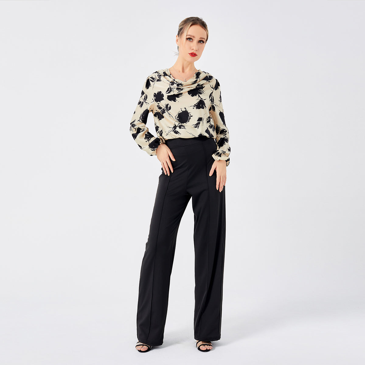 Elegante Frau in schwarz-weißer Bluse mit Blumenmuster und schwarzer Hose, posiert vor weißem Hintergrund. Modetrend, Damenbekleidung, stilvoll.