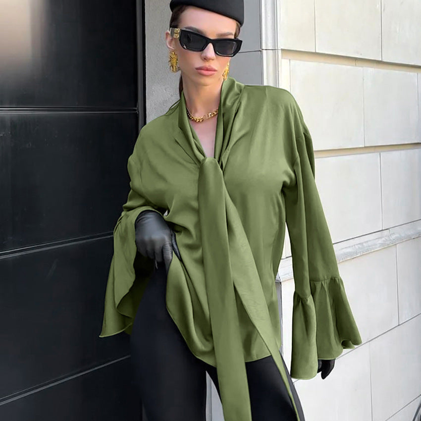 Frau in grüner Bluse mit Krawattenkragen, schwarzer Hose und Sonnenbrille, posiert vor moderner Architektur. Elegante Mode, urbaner Stil.