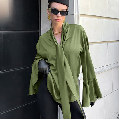 Frau in grüner Bluse mit Krawattenkragen, schwarzer Hose und Sonnenbrille, posiert vor moderner Architektur. Elegante Mode, urbaner Stil.