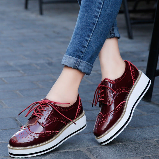 Damenbeine in blauen Jeans tragen rote Lackleder-Brogues mit Plateauabsatz auf gepflastertem Boden. Modische Schuhe, trendiger Look.