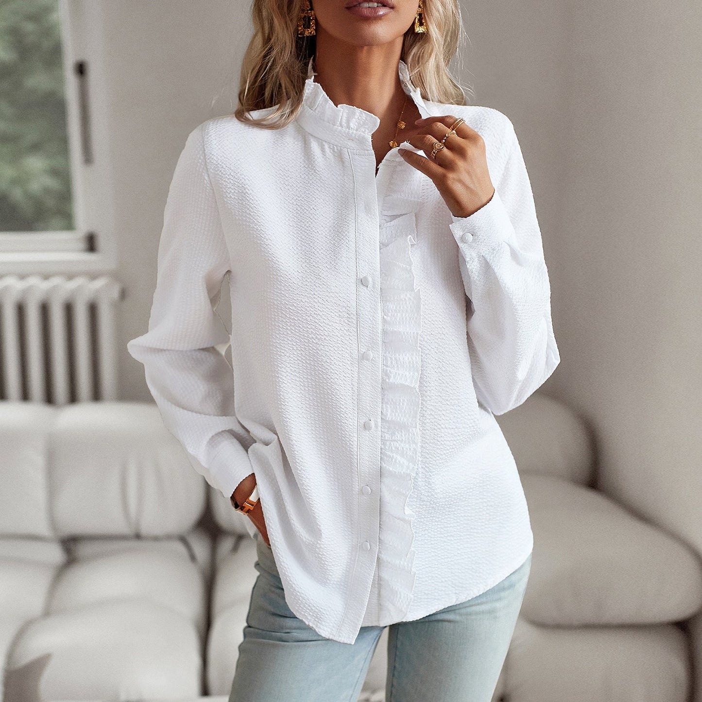 Frau in weißer Bluse mit Rüschen, steht in modernem Wohnzimmer. Elegante Mode, lässiger Stil, helle Jeans. Perfekt für stilvolle Outfits.