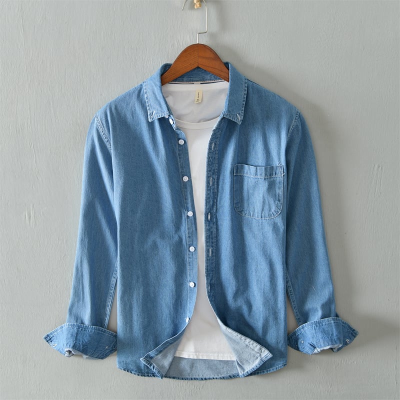 Hellblaues Jeanshemd auf Holzkleiderbügel, über weißem T-Shirt, vor grauer Wand. Modetrend, Freizeitkleidung, Denim-Look.