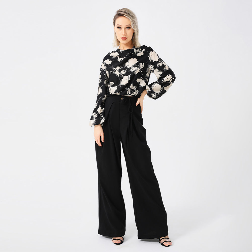 Frau in eleganter, schwarzer Hose und geblümter Bluse, posiert vor weißem Hintergrund. Modetrend, Damenbekleidung, stilvolle Outfits.