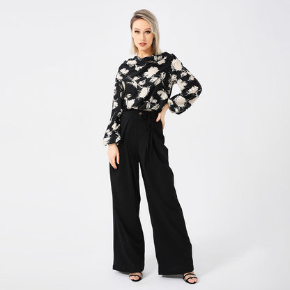 Frau in eleganter, schwarzer Hose und geblümter Bluse, posiert vor weißem Hintergrund. Modetrend, Damenbekleidung, stilvolle Outfits.