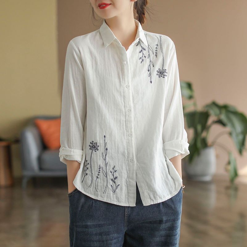 Frau trägt weiße Bluse mit floraler Stickerei, lässig gekrempelte Ärmel, kombiniert mit blauen Jeans. Elegante Freizeitmode, modernes Design.
