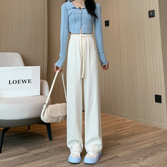 Frau in blauem Oberteil und weißen Hosen posiert in modernem Raum mit Loewe-Tasche. Modetrend, stilvolle Damenbekleidung, elegantes Outfit.