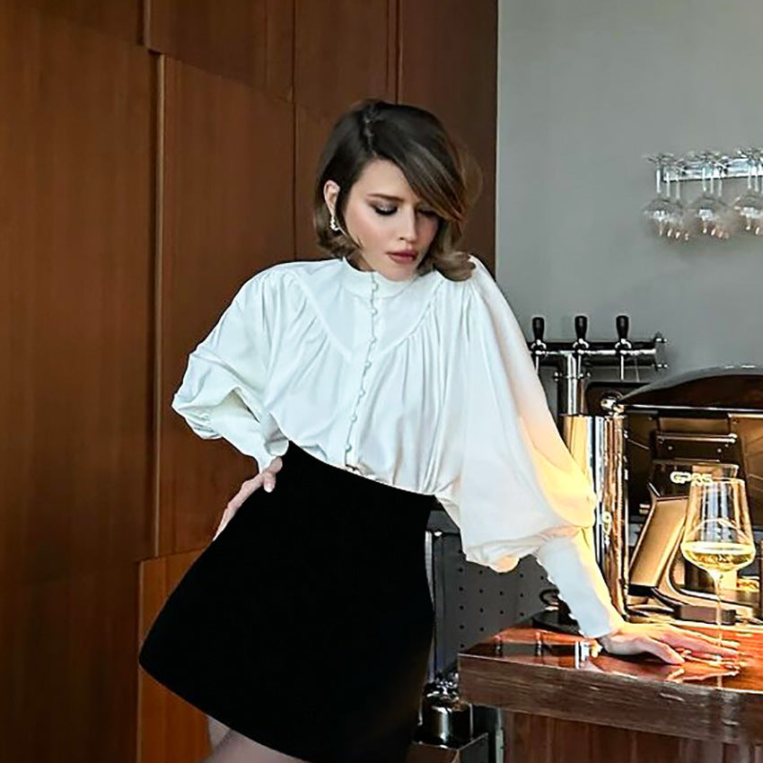 Frau in stilvoller weißer Bluse und schwarzem Rock lehnt an Bartheke mit Weinglas, moderne Inneneinrichtung, elegante Mode, Abendgarderobe.