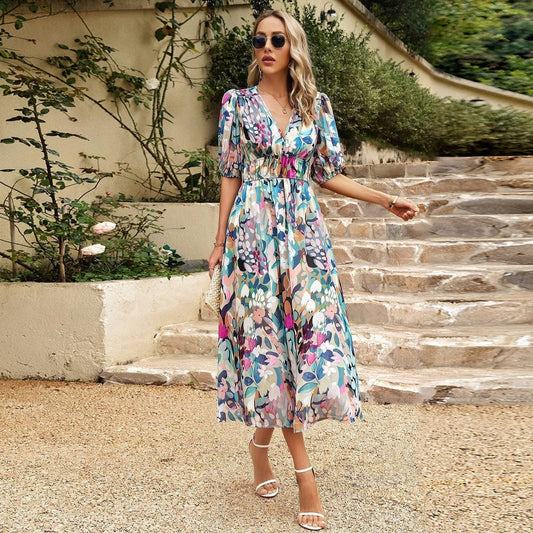 Frau in geblümtem Sommerkleid auf Steintreppe, trägt Sonnenbrille und Sandalen, umgeben von grüner Vegetation. Elegante Mode, Outdoor-Stil.
