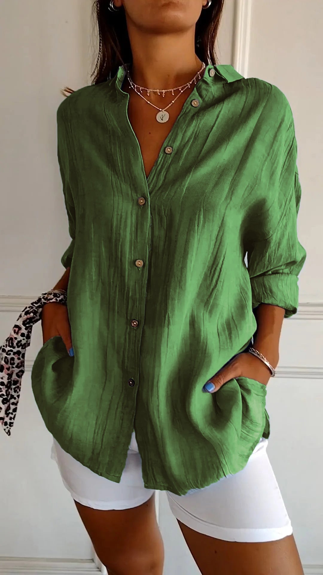 Frau trägt grüne Leinenbluse mit Knopfleiste und weißen Hosen. Modischer Look, ideal für Sommer. Accessoires: Halskette, Armband, gemusterter Schal.
