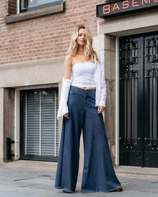Frau in weißem Top und blauer Schlaghose vor Backsteingebäude mit "Basement"-Schild. Modetrend, lässiger Streetstyle, urbane Mode.