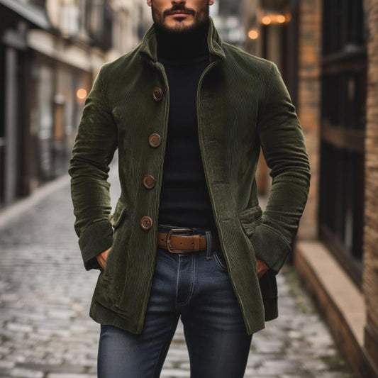 Mann in grünem Cordmantel und schwarzem Rollkragenpullover auf Kopfsteinpflasterstraße. Modischer Herbstlook, stilvolle Herrenmode, urbane Kulisse.